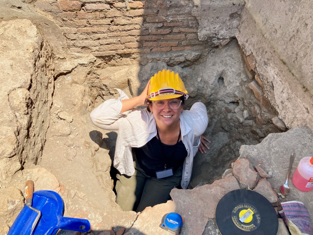 A smiling woman wearing a yellow hard hat is sitting in an archaeological excavation site, surrounded by rocky soil and brick walls.
